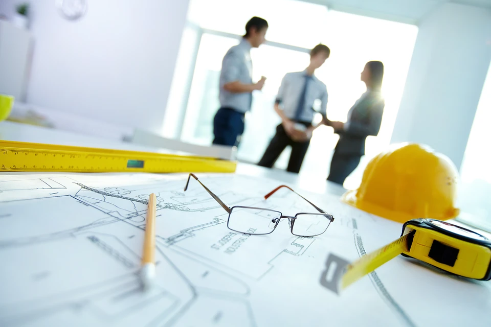Architecte ou ingénieur en chemise à carreaux travaillant sur des plans techniques dans un bureau lumineux avec grande fenêtre donnant sur verdure